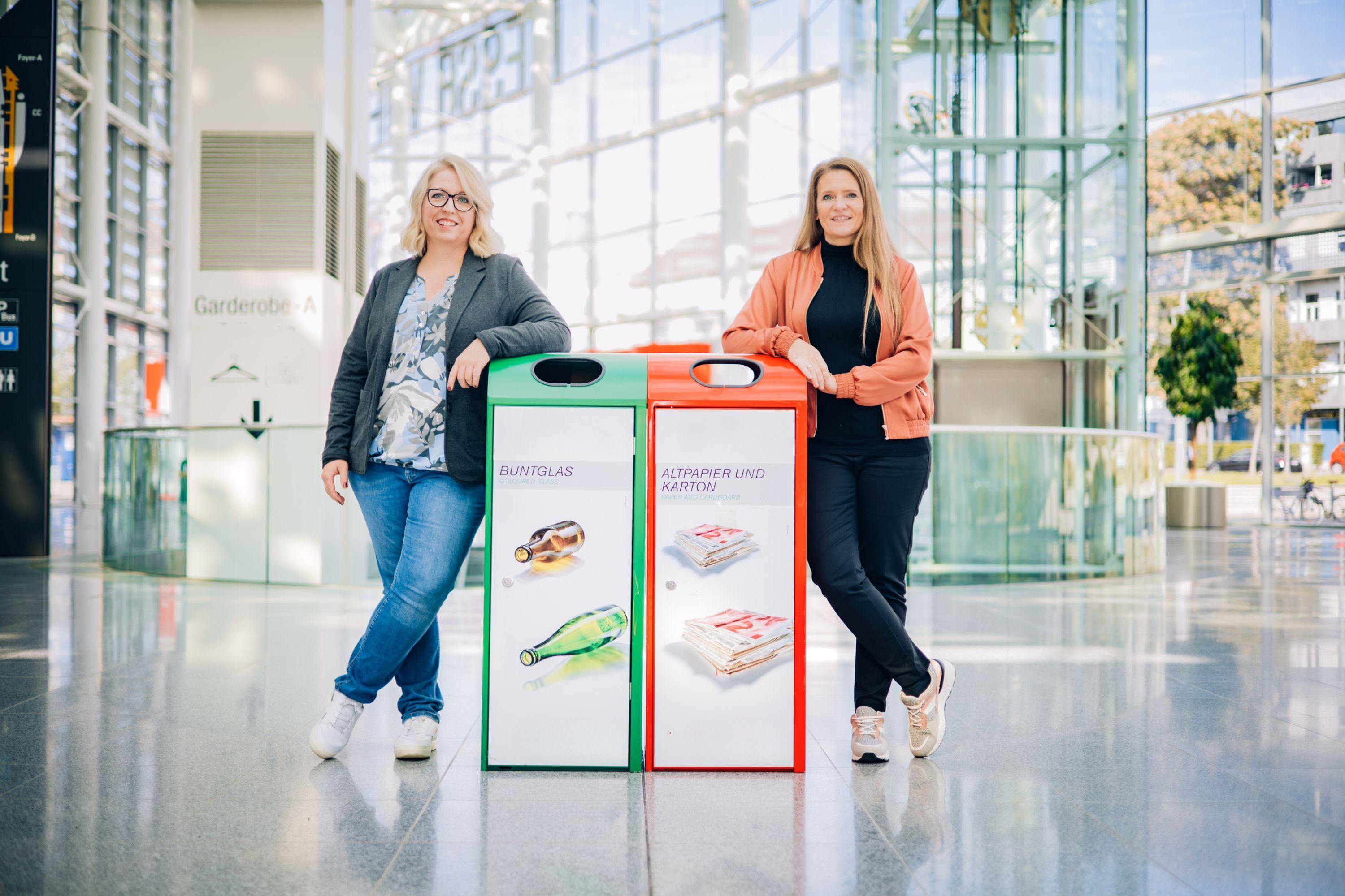 Susanne Zehntner, Head of Sustainability, und Silvia Hiller, Technical Administration Manager blicken in die Kamera und lehnen sich an den Containern zur Mülltrennung an.