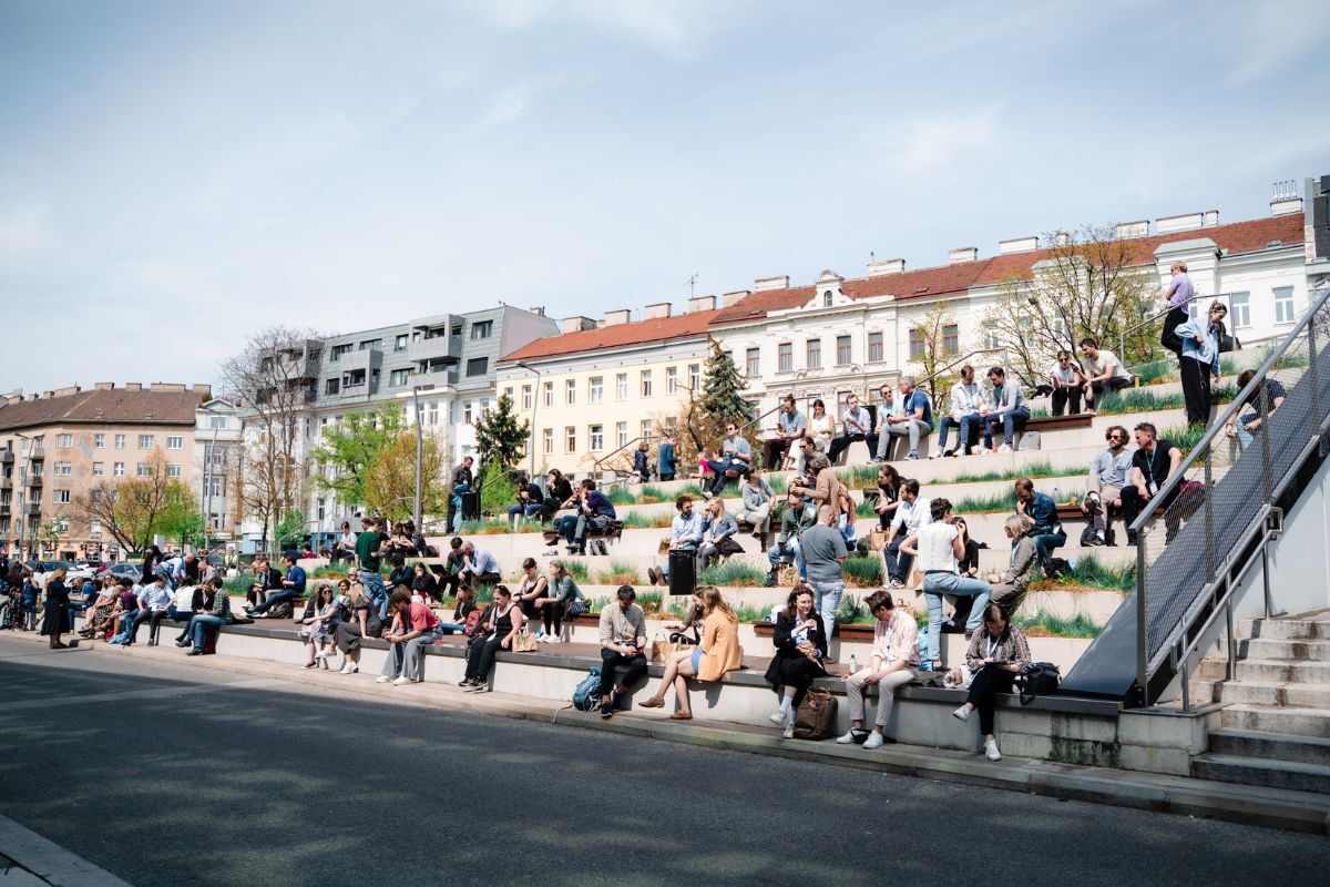 Menschen sitzen und entspannen sich an einem sonnigen Tag auf einer Freitreppe in einer lebhaften städtischen Umgebung neben der VIECON.